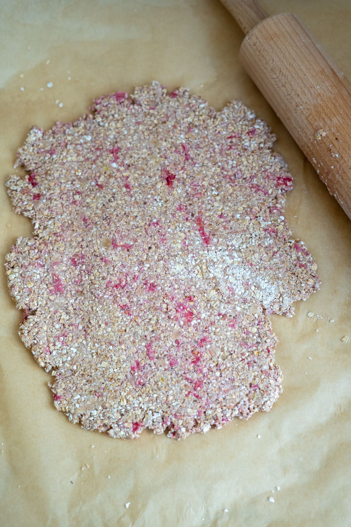 Porridge and berry dough rolled out on baking parchment next to a wooden rolling pin.