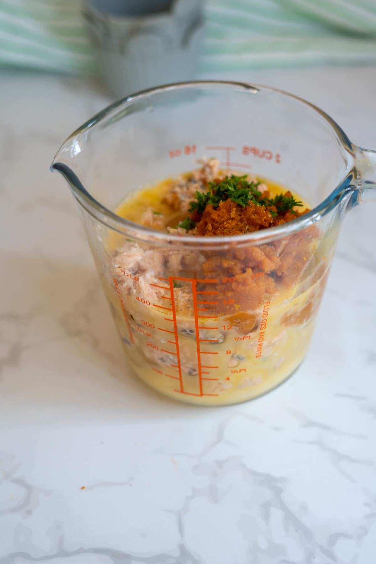 Glass measuring jug with eggs, tuna, breadcrumbs, and chopped parsley on a white marble worktop.