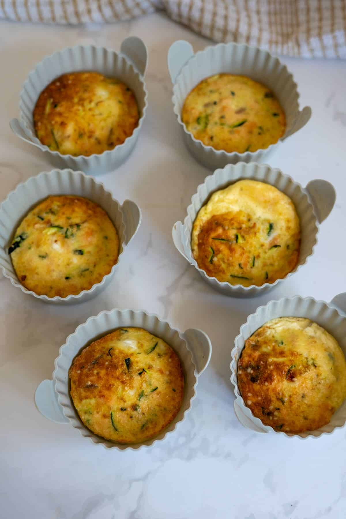 Six baked muffins with visible green herbs are in light grey silicone baking cups on a white surface, reminiscent of a savoury Air Fryer Salmon Dog Frittata.