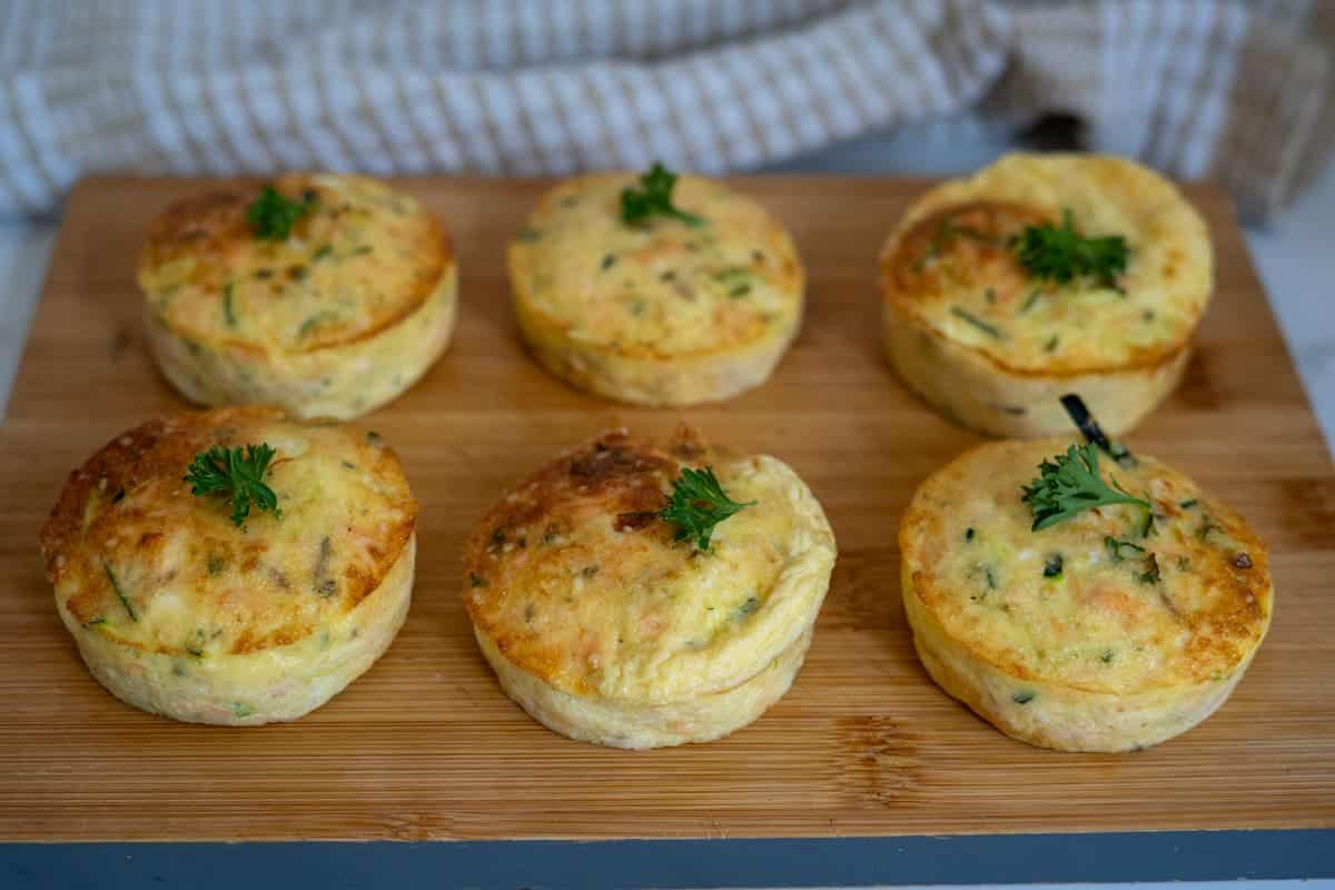 Six baked egg muffins with herbs on top are arranged on a wooden chopping board, reminiscent of a mini frittata—perfect for pairing with your favourite Air Fryer Salmon Dog or serving as a wholesome breakfast option. A striped tea towel in the background adds a cosy touch.