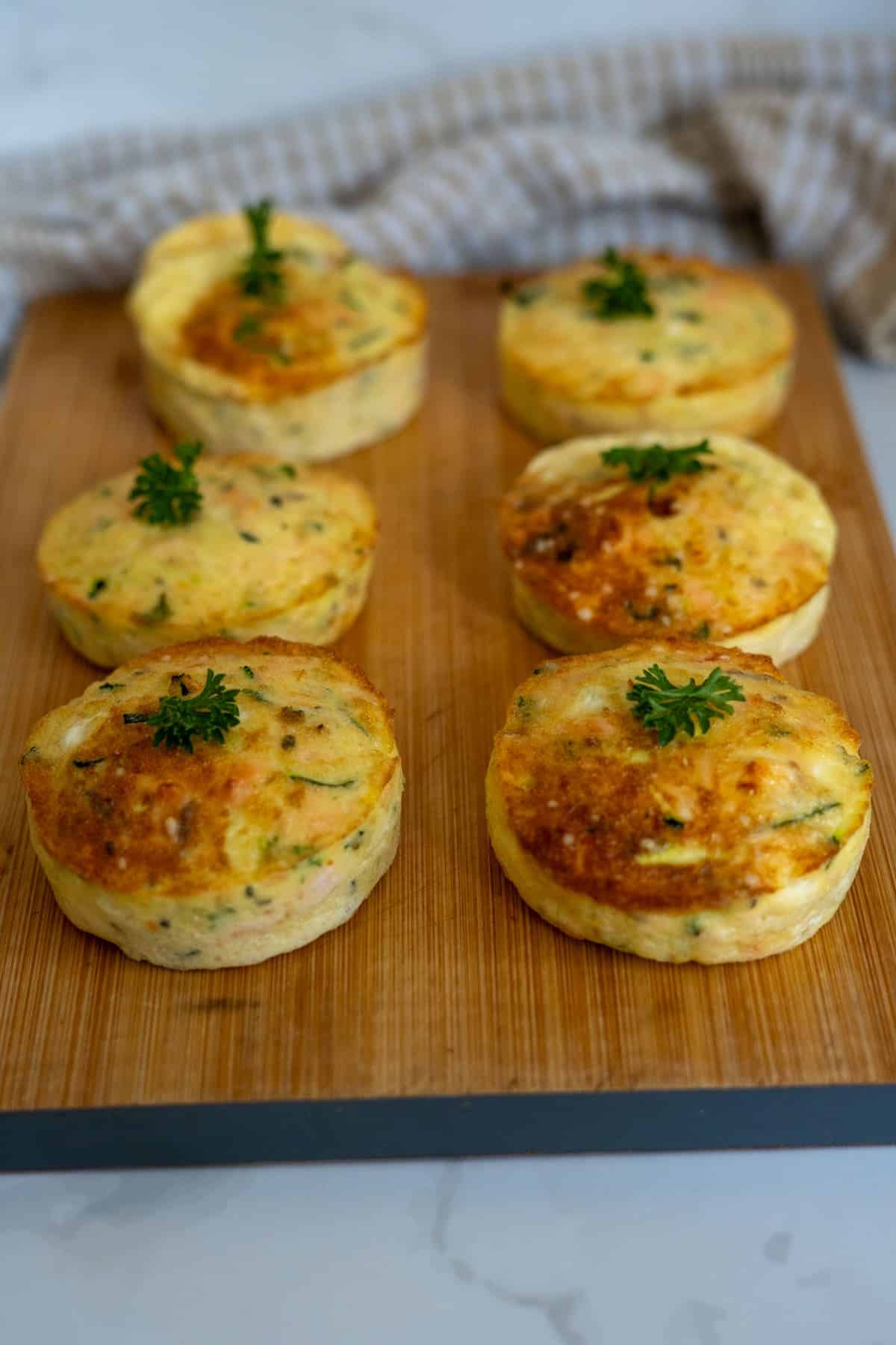 Six baked egg muffins with herbs on top are arranged on a wooden chopping board, reminiscent of a savoury Air Fryer Salmon Dog Frittata. A light-coloured cloth is visible in the background.