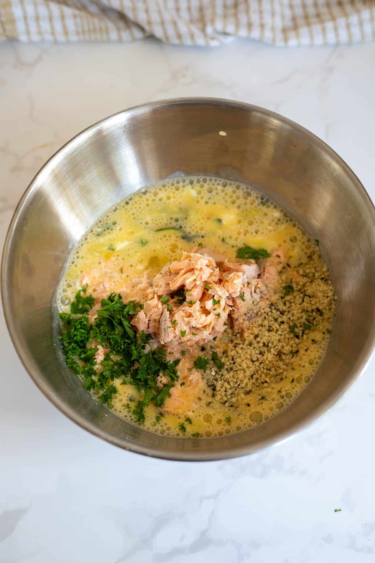 A metal bowl containing beaten eggs, flaked salmon, chopped parsley, and breadcrumbs on a white worktop—perfect ingredients for whipping up an Air Fryer Salmon Dog Frittata.