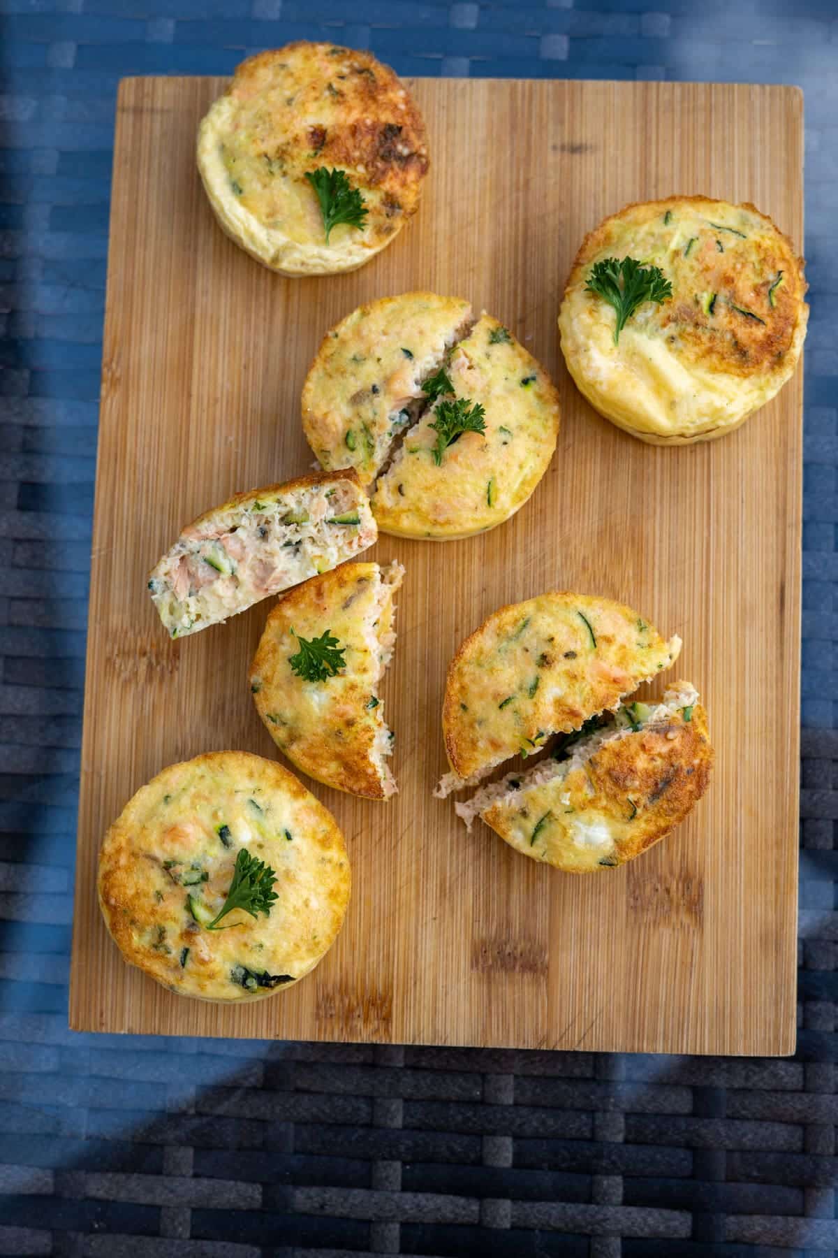 Six egg muffins with herbs, reminiscent of a mini Air Fryer Salmon Dog Frittata, are arranged on a wooden board. Some are cut in half to reveal the savoury filling inside, and each muffin is garnished with a small parsley leaf.