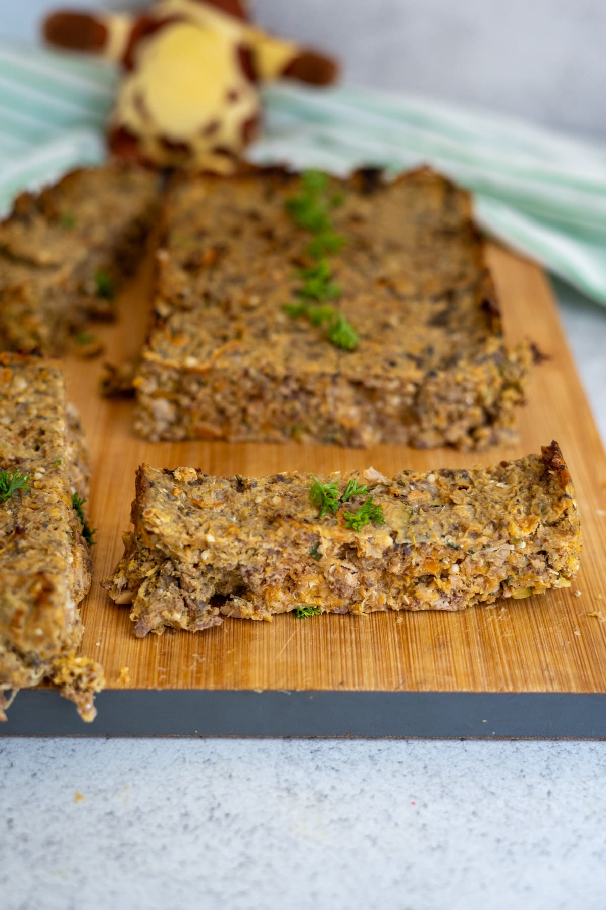 A loaf of vegetable and nut roast is sliced on a wooden board, garnished with green herbs.