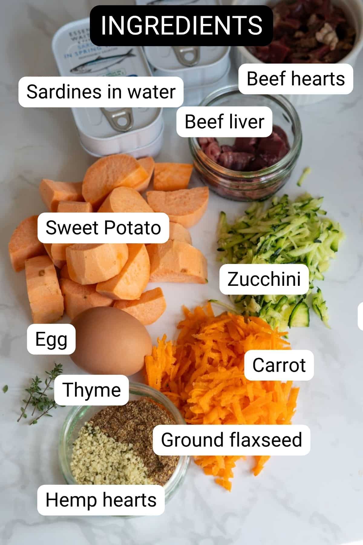 Various labelled ingredients including sardines in water, beef hearts, beef liver, sweet potato, courgette, carrot, egg, thyme, ground linseed, and hemp hearts are arranged on a counter.