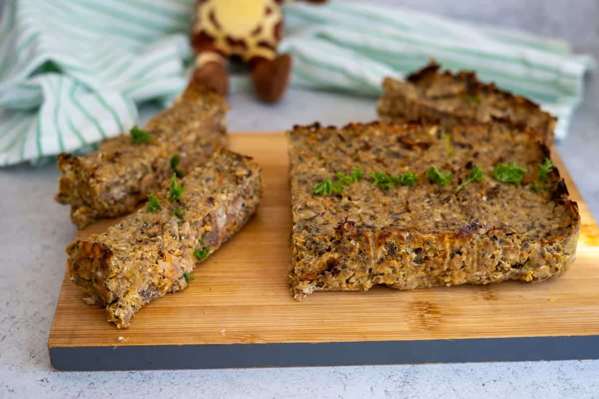 A rectangular loaf with three slices cut, garnished with herbs, sits on a wooden chopping board; a striped cloth and soft toy are in the background.