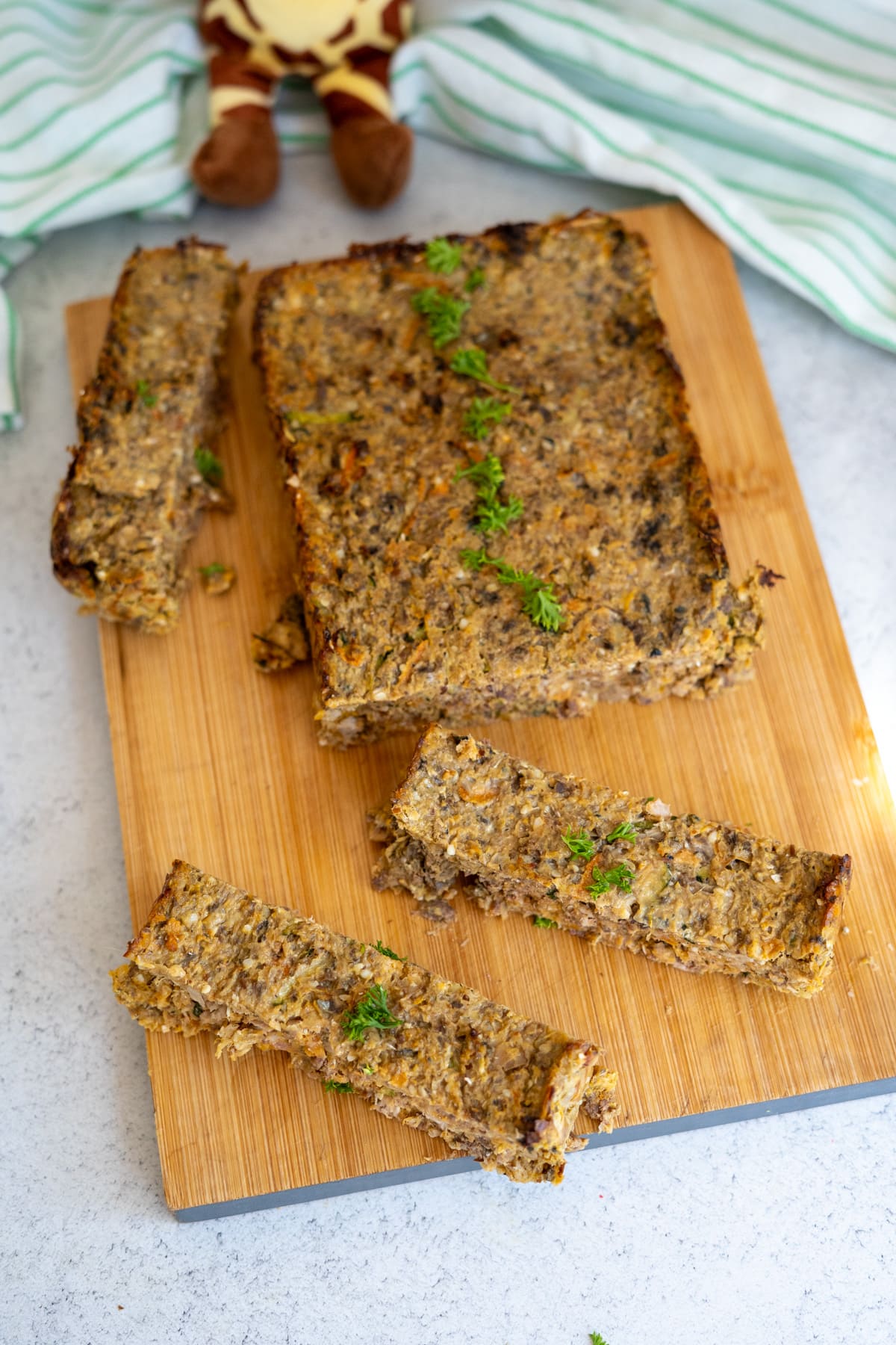 A rectangular loaf of vegetable and grain mixture is sliced on a wooden chopping board, garnished with fresh herbs.