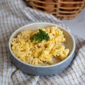A plate of scrambled eggs garnished with a sprig of parsley sits on a striped cloth with a basket in the background.