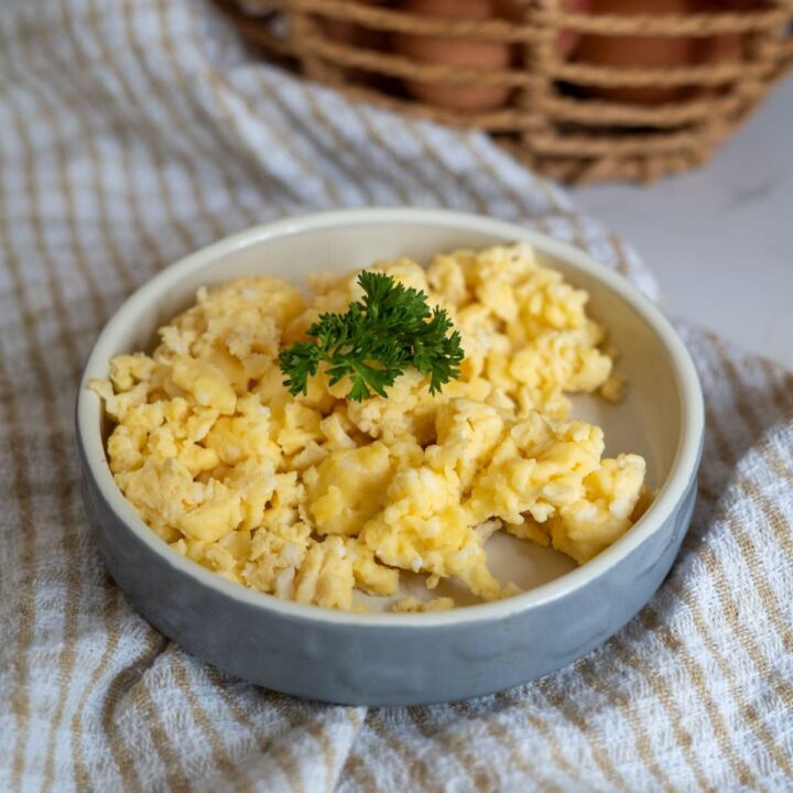A plate of scrambled eggs garnished with a sprig of parsley sits on a striped cloth with a basket in the background.