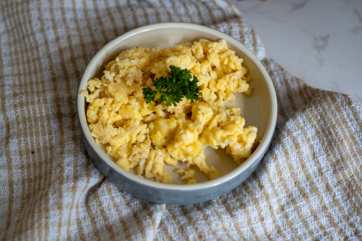 Scrambled eggs for dogs served in a round bowl, garnished with a sprig of parsley, placed on a beige and white checked cloth.