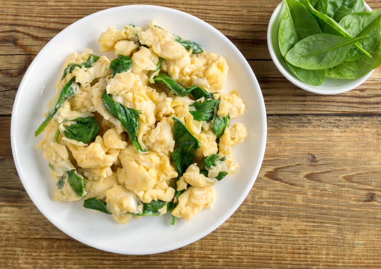 A plate of scrambled eggs with spinach sits next to a small bowl of fresh spinach leaves on a wooden surface, prompting the question: can dogs eat scrambled eggs?.