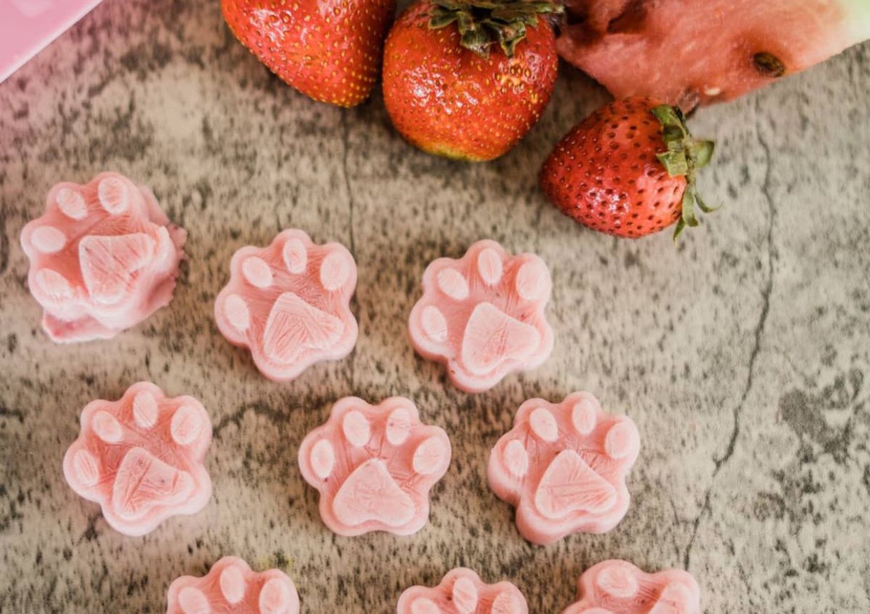 Pink paw-shaped treats are arranged on a grey surface near whole strawberries and a slice of watermelon, offering a charming example of creative strawberry dog treat recipes.