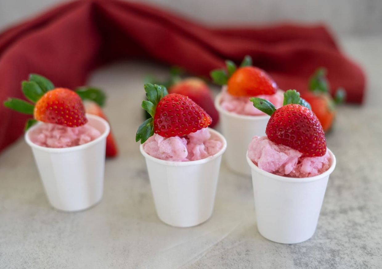 Five small white cups filled with pink strawberry ice cream, each topped with a whole strawberry, are arranged on a light surface with a red cloth in the background, prompting the question: can dogs eat strawberry ice cream?.