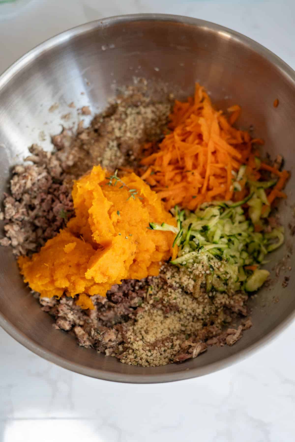 A metal bowl containing minced meat, grated carrots, grated courgette, hemp seeds, and mashed sweet potatoes on a white surface.