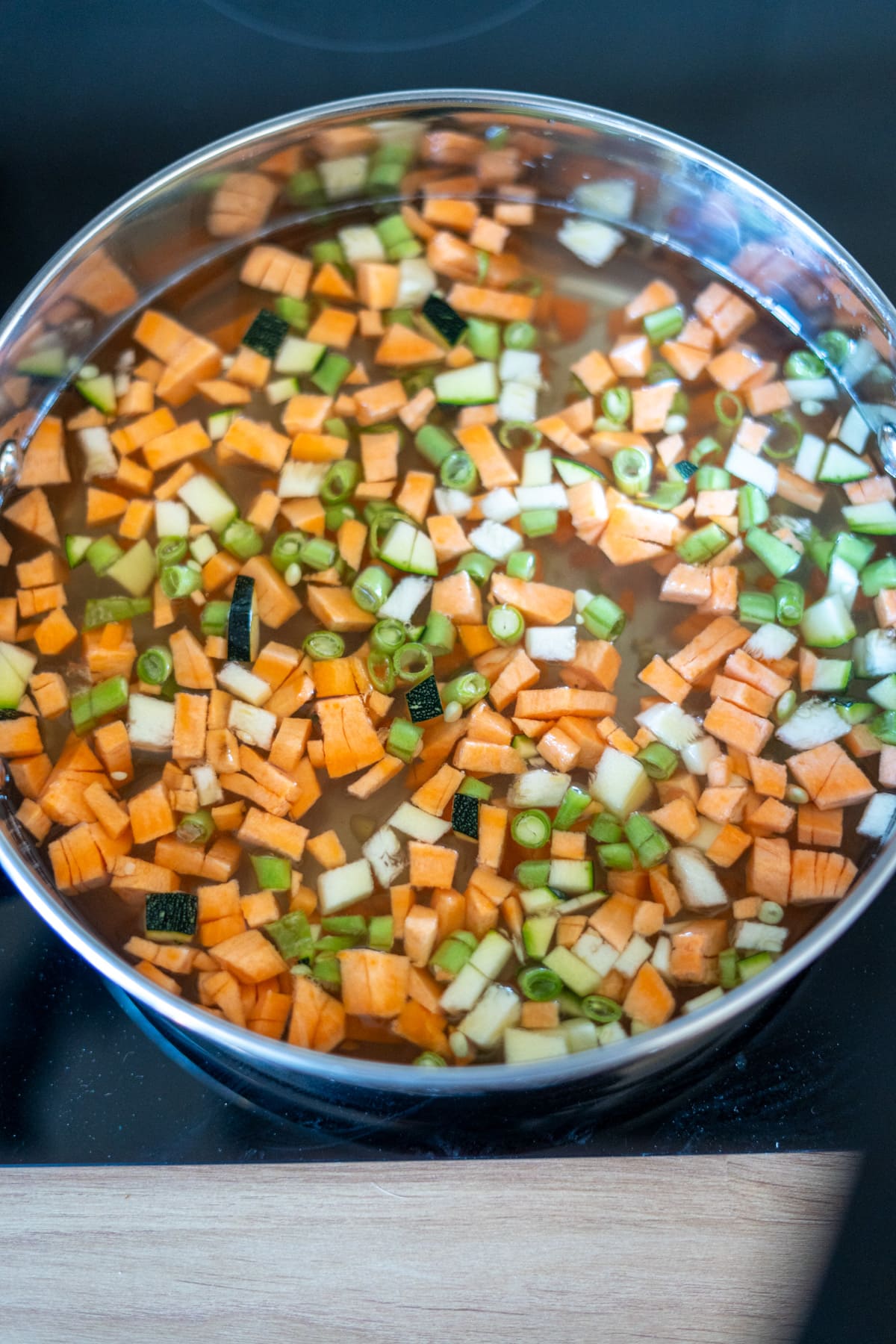 A pot filled with water and diced vegetables, including sweet potatoes, courgettes, and spring onions, simmers on a hob as the perfect base for a nourishing fish stew for dogs.