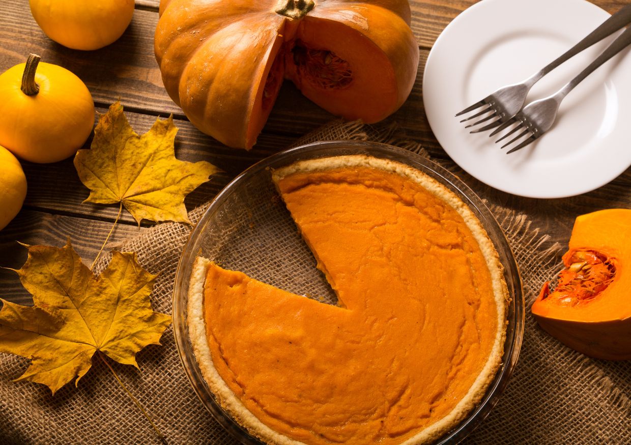 A pumpkin pie with one slice missing sits on a wooden table alongside pumpkins, autumn leaves, a plate, and two forks.