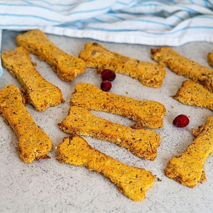 Bone-shaped Turkey Pumpkin Cranberry Dog Treats are arranged on a light surface with three red cranberries nearby; a striped cloth is in the background.