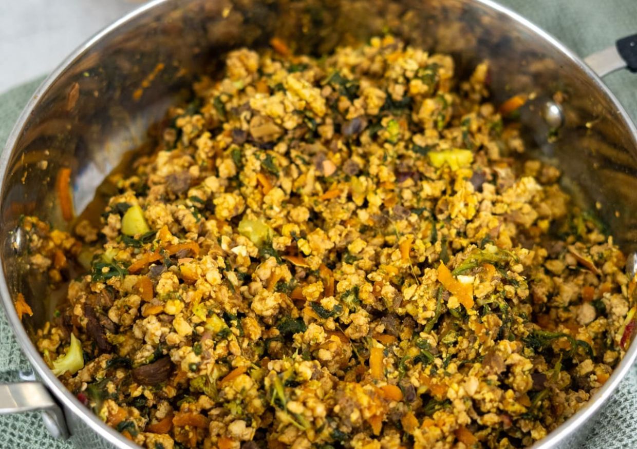 A large metal pan filled with a cooked mixture of crumbled tofu, vegetables, and seasonings on a green cloth—similar to recipes you’ll find when learning how to cook minced turkey for dogs, but made entirely plant-based.