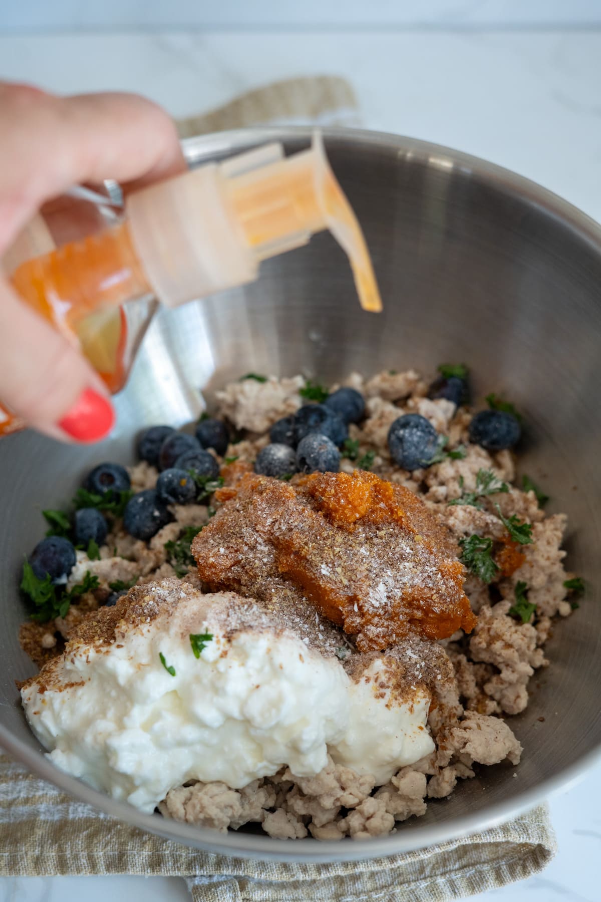 A hand presses a pump bottle over a metal bowl filled with minced meat, blueberries, cottage cheese, herbs, and spices.