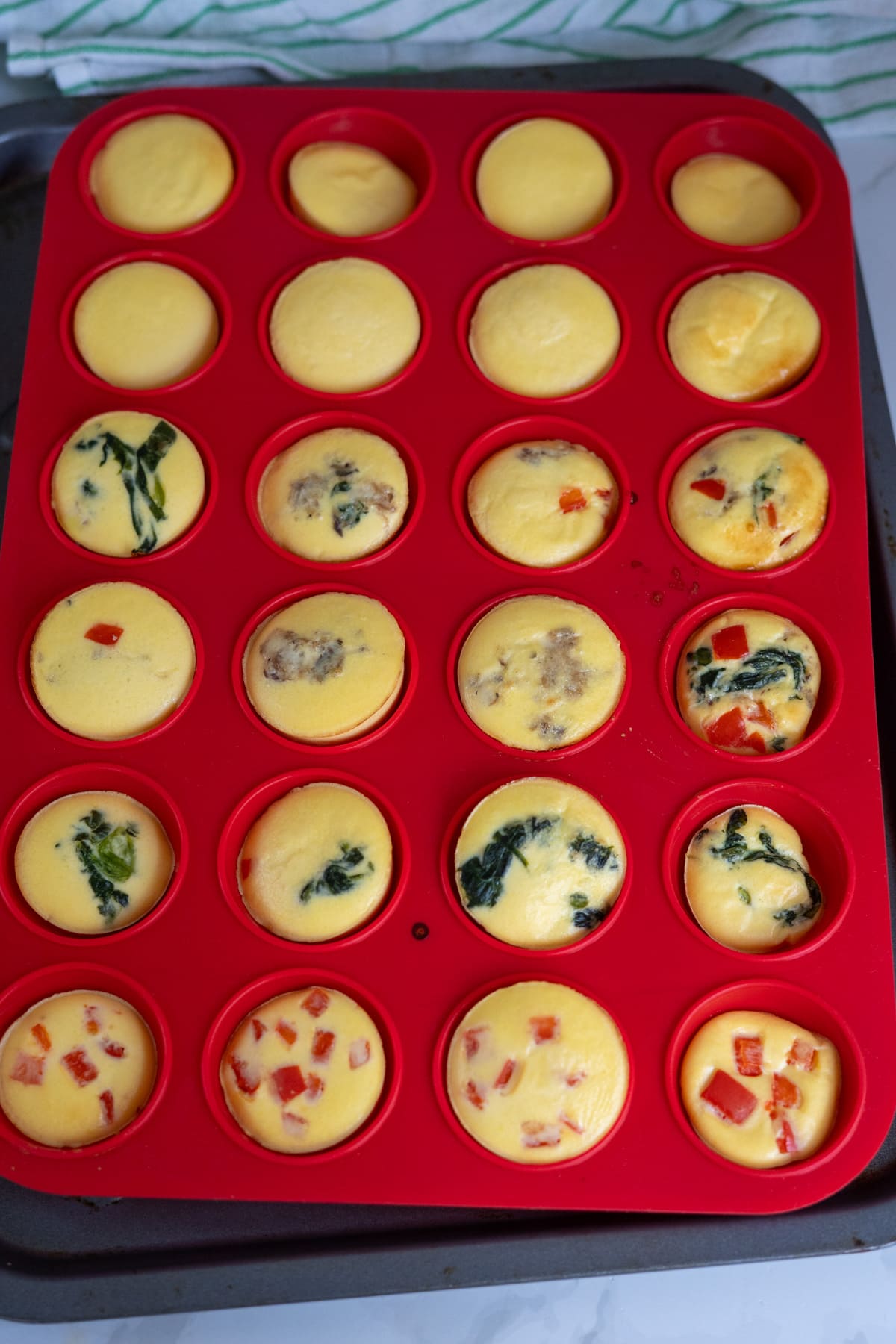 A red silicone muffin tray holds 24 baked egg bites, some with spinach, sausage, or red pepper, on a baking tray—perfect for making nutritious egg bites for dogs.