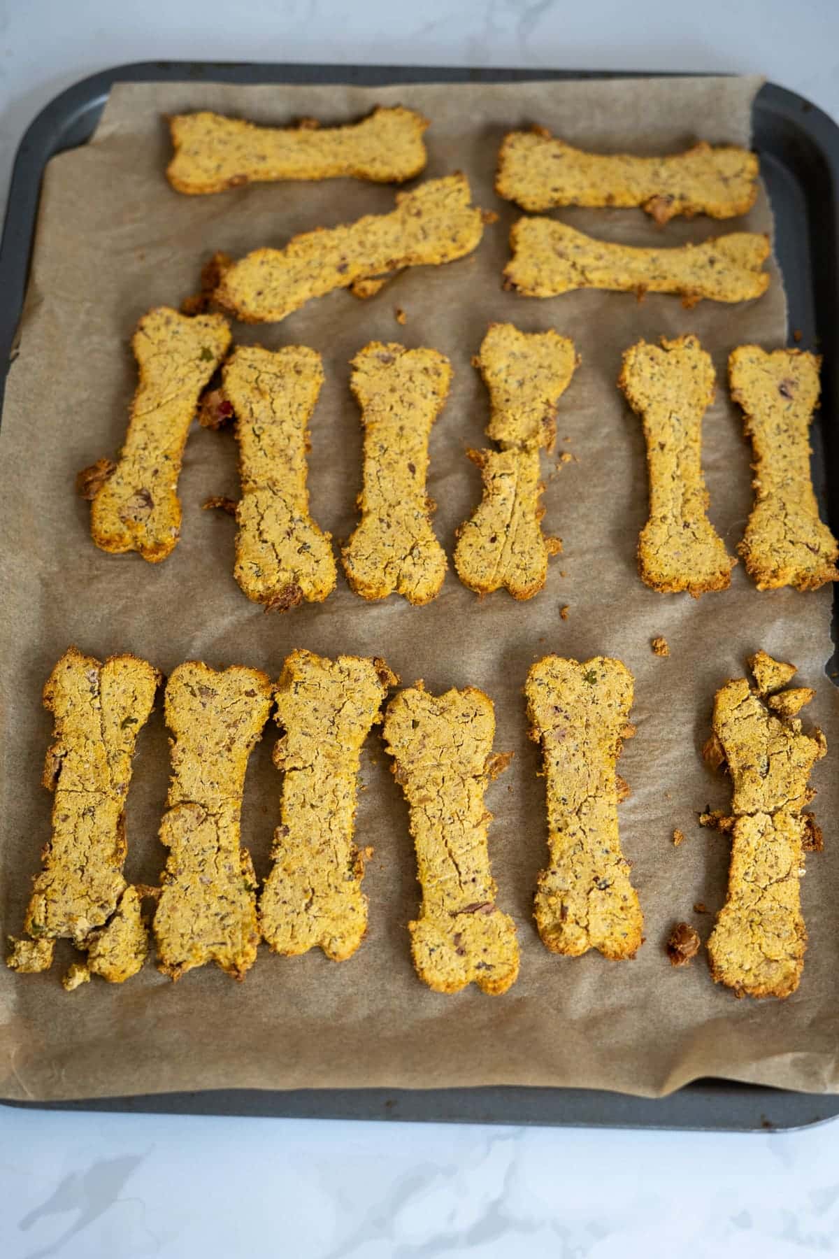 A baking tray lined with greaseproof paper holds several bone-shaped Turkey Pumpkin Cranberry Dog Treats arranged in rows.