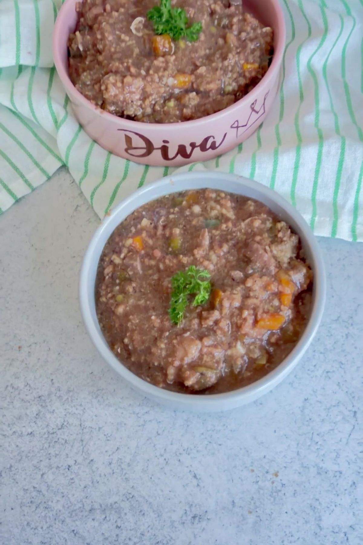 Two bowls of chunky stew with vegetables, garnished with parsley, sit on a light grey surface with a striped cloth in the background, resembling a delicious homemade Slow Cooker Beef Dog Food recipe.