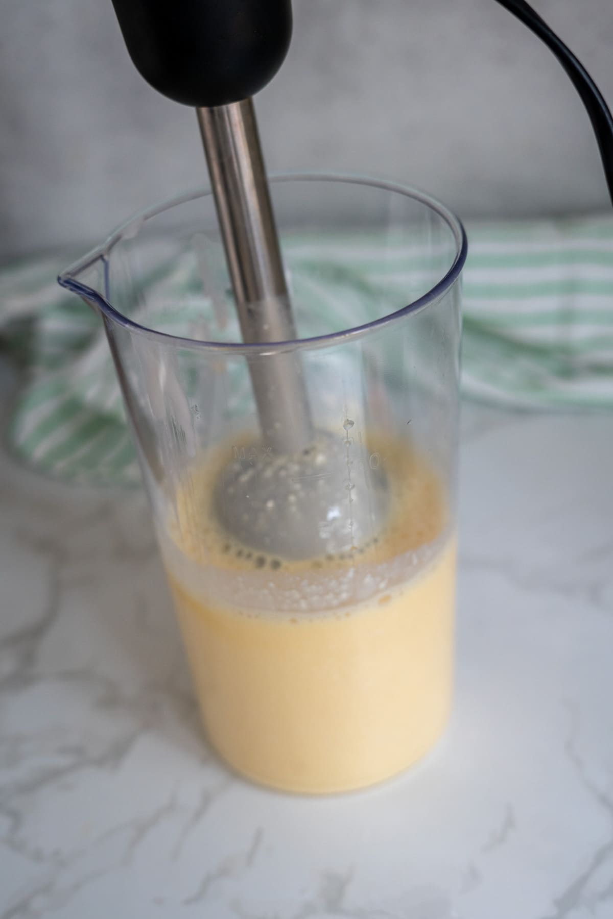 A hand blender is mixing a yellow liquid for egg bites for dogs in a clear plastic container on a marble worktop, with a green and white striped cloth in the background.