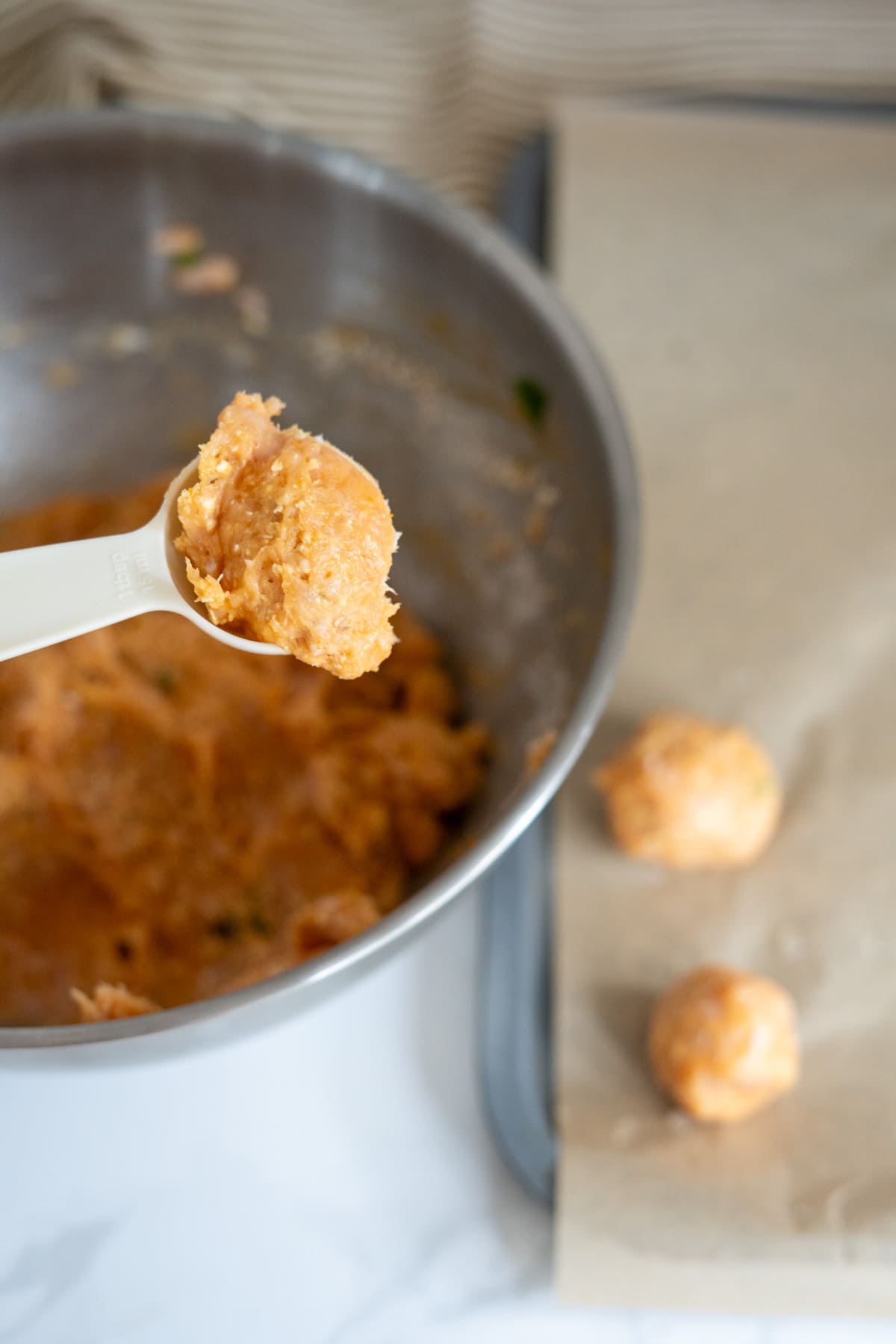 A spoon holds raw mince mixture above a bowl, with two formed meatballs resting on a baking tray lined with baking parchment beside it.