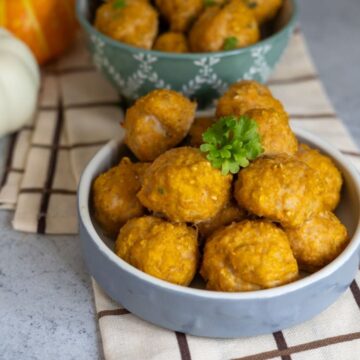 Two bowls filled with round, golden-brown cheesy snacks are placed on a chequered cloth, with a sprig of parsley on top for garnish.