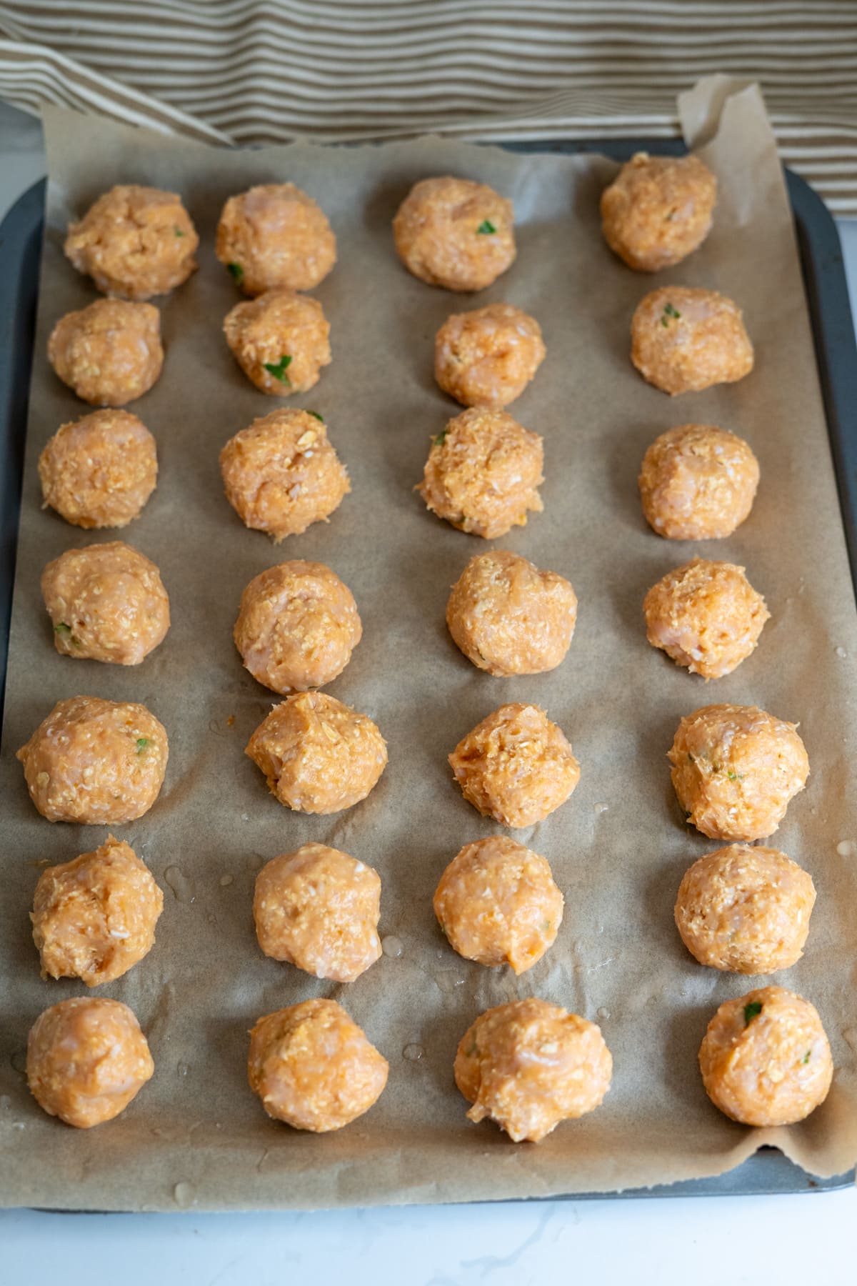 A baking tray lined with greaseproof paper holds five rows of uncooked meatballs evenly spaced in a grid pattern, ready for baking.