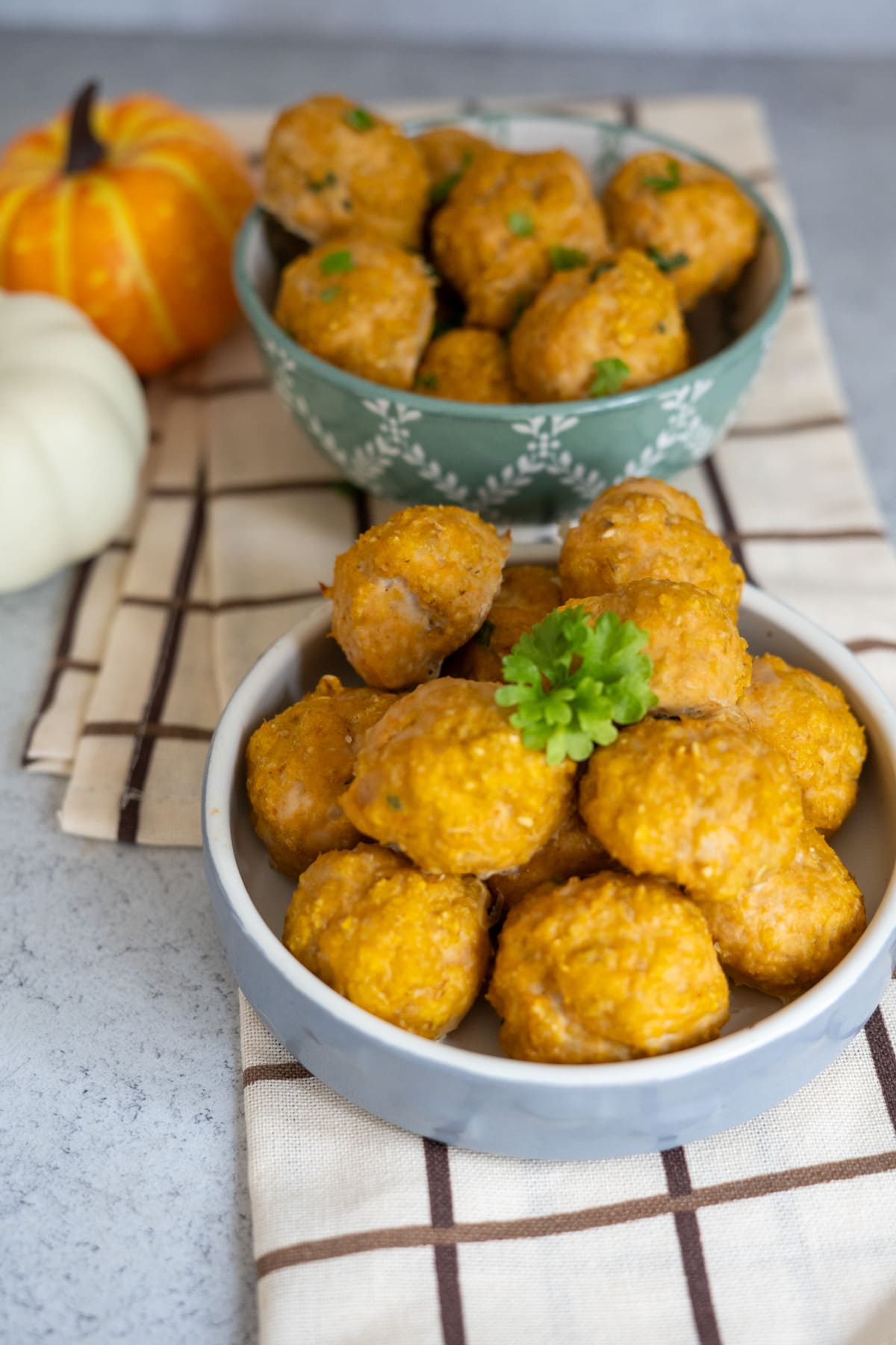 Two bowls filled with round, golden-brown chicken meatballs for dogs are placed on a chequered cloth, with two decorative pumpkins in the background.