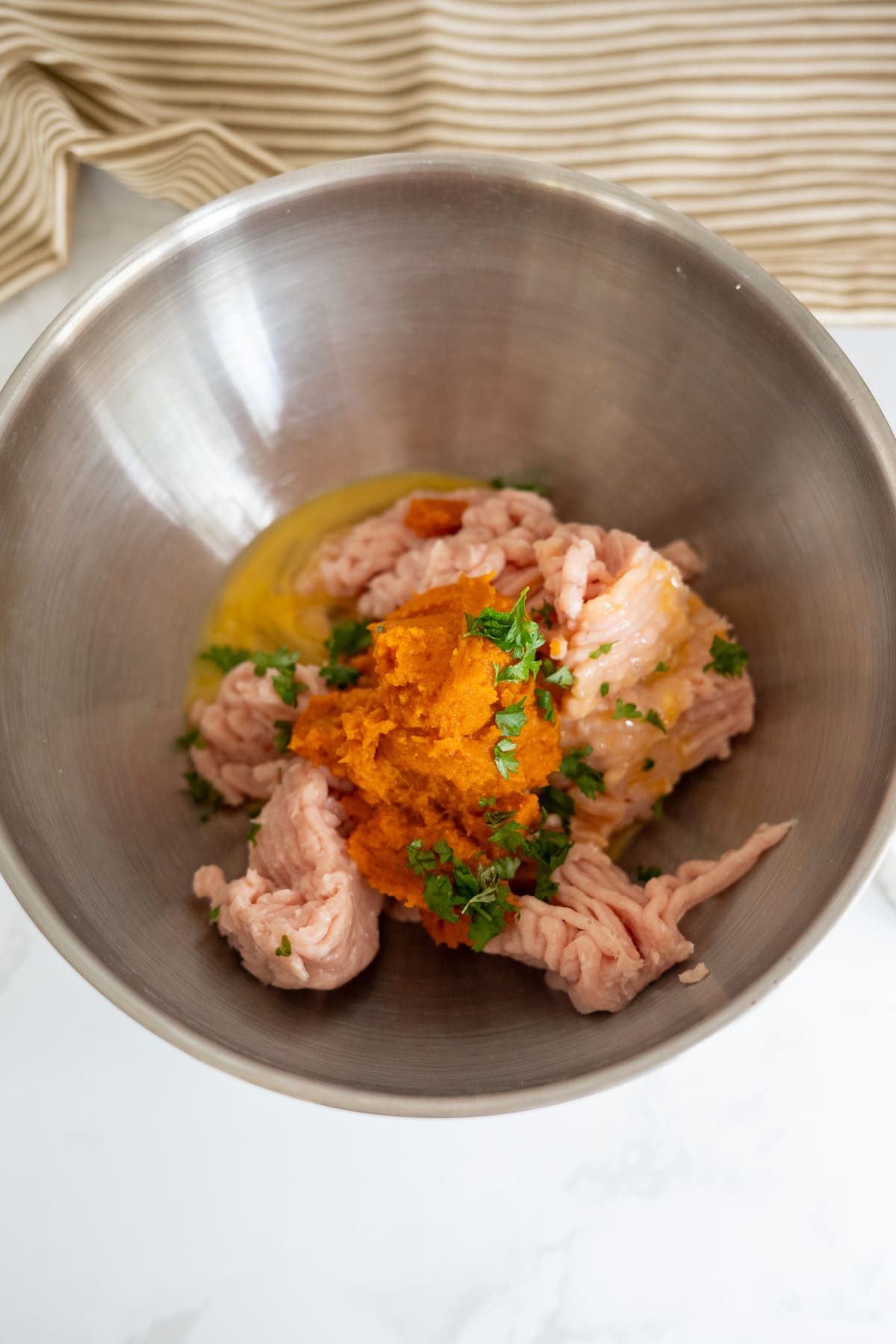 Minced meat, mashed sweet potato, chopped herbs, and an egg yolk in a large metal mixing bowl on a white worktop.