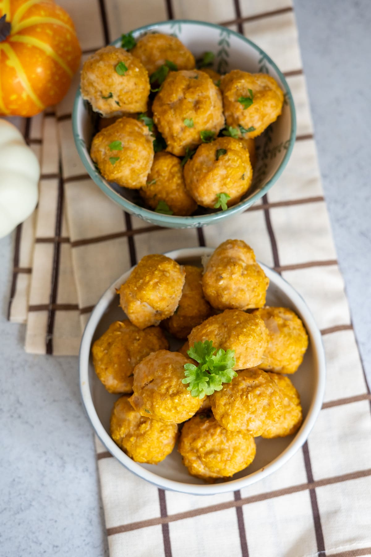 Two bowls of pumpkin-coloured chicken meatballs for dogs are placed on a striped cloth, garnished with green herbs, with small decorative pumpkins nearby.