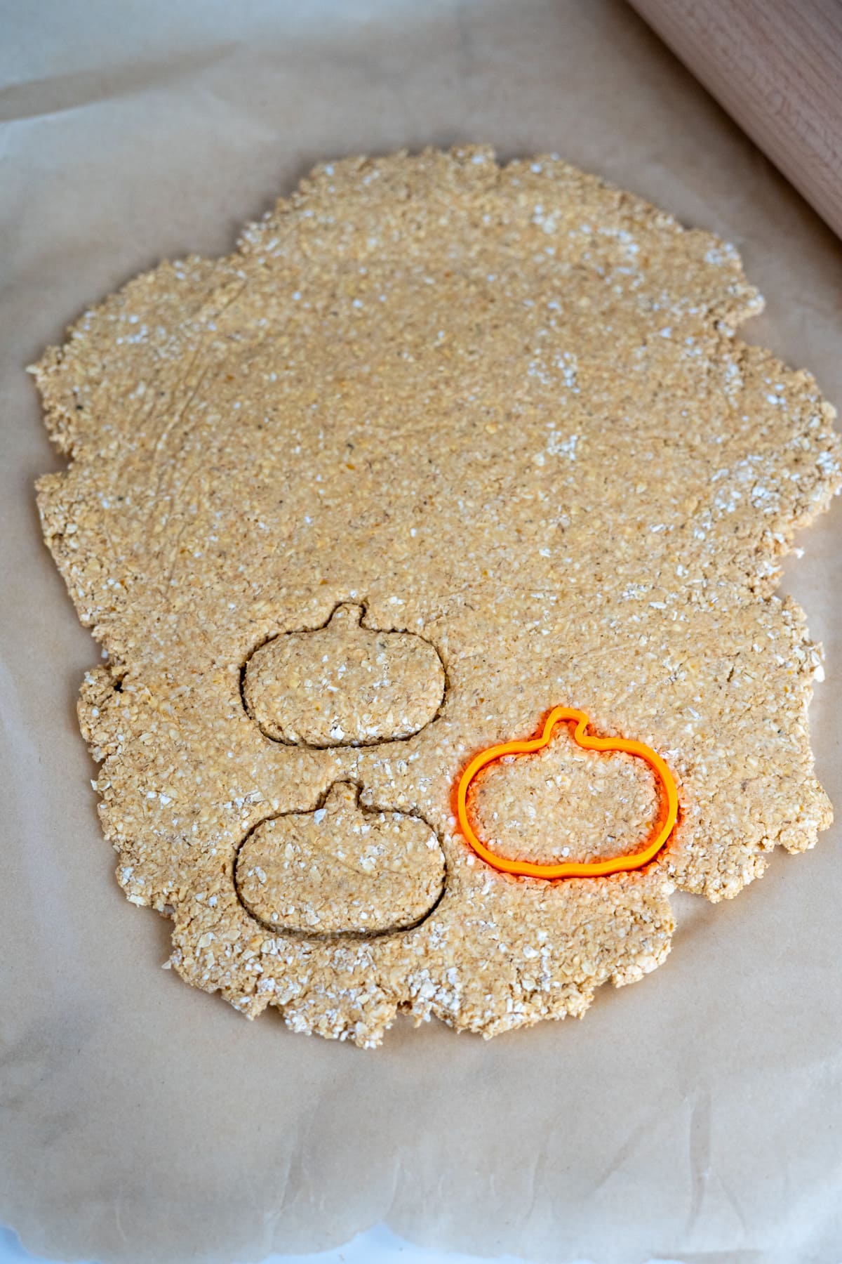 Pumpkin-shaped biscuit cutter pressed into rolled-out dough on baking paper, with two pumpkin outlines visible nearby—perfect for making homemade goat milk pumpkin dog biscuits.
