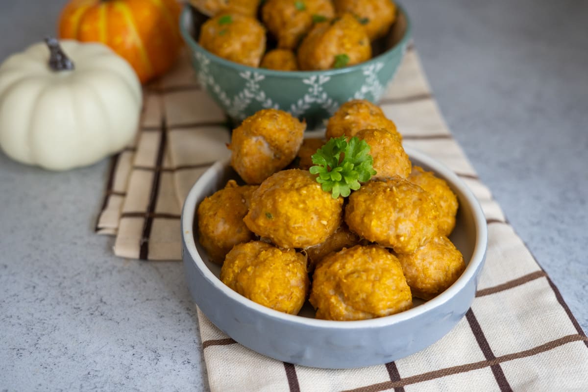Two bowls of round, golden-brown snacks are on a chequered cloth, with decorative pumpkins in the background and a sprig of parsley on top of the front bowl.