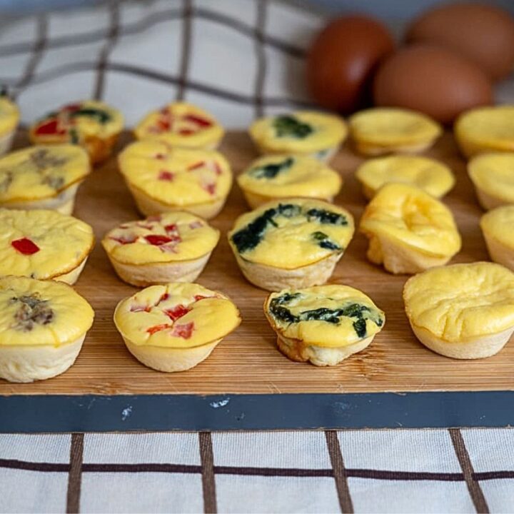 Mini quiches with various fillings are arranged on a wooden board. Three brown eggs are in the background, and the board is on a chequered tablecloth.