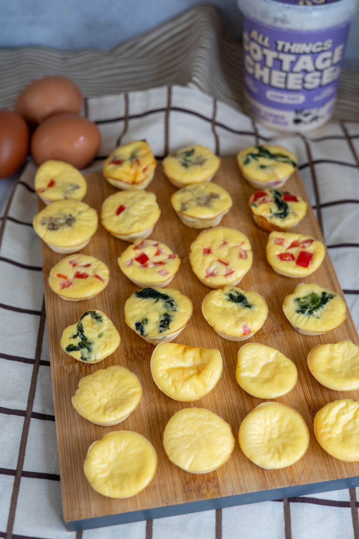 Mini egg bites for dogs with various fillings are arranged on a wooden board. Three brown eggs and a tub of cottage cheese are in the background on a checked cloth.