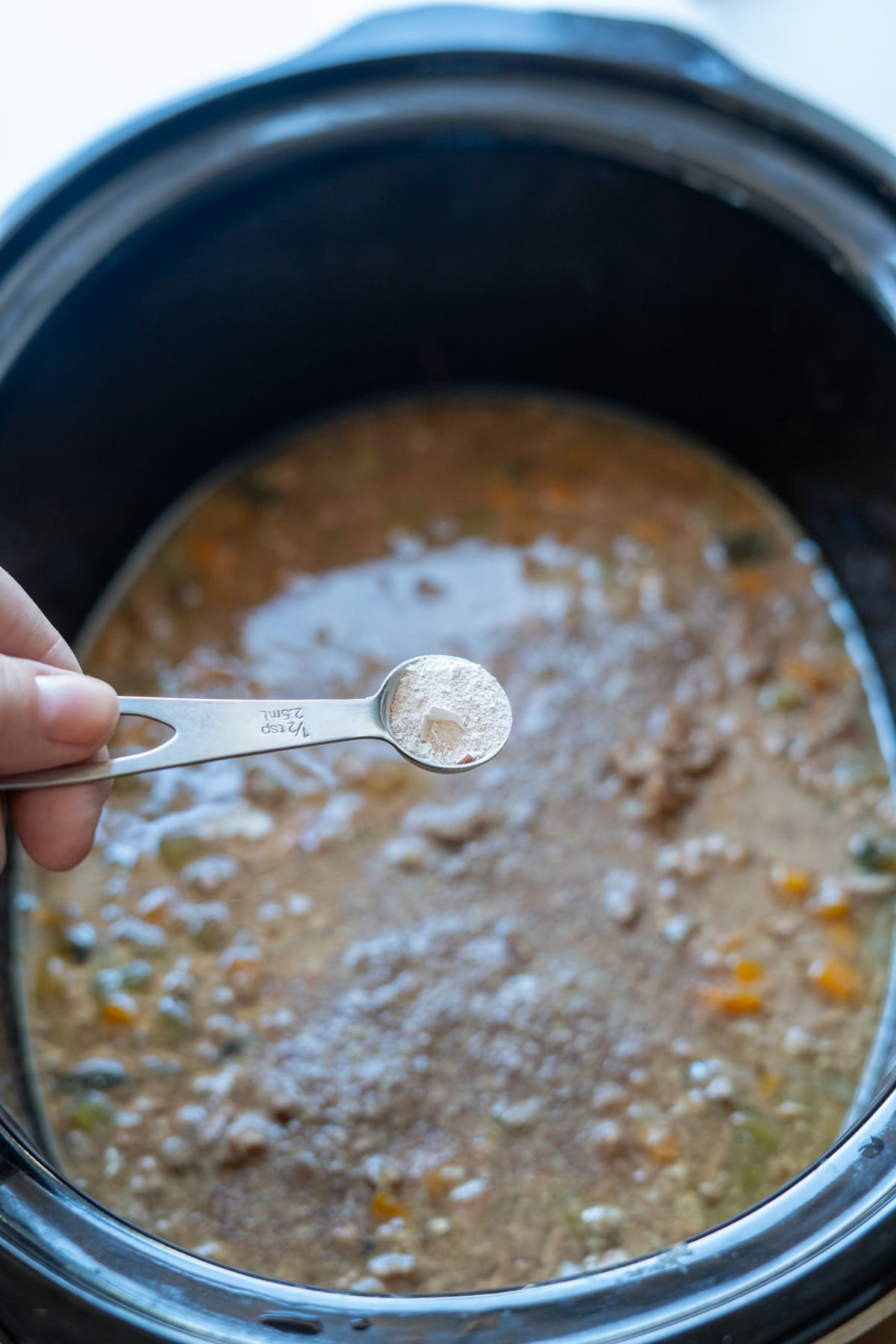 A hand holds a measuring spoon with flour over a slow cooker containing a mixture of stock, vegetables, and meat, as part of preparing homemade Slow Cooker Beef Dog Food.