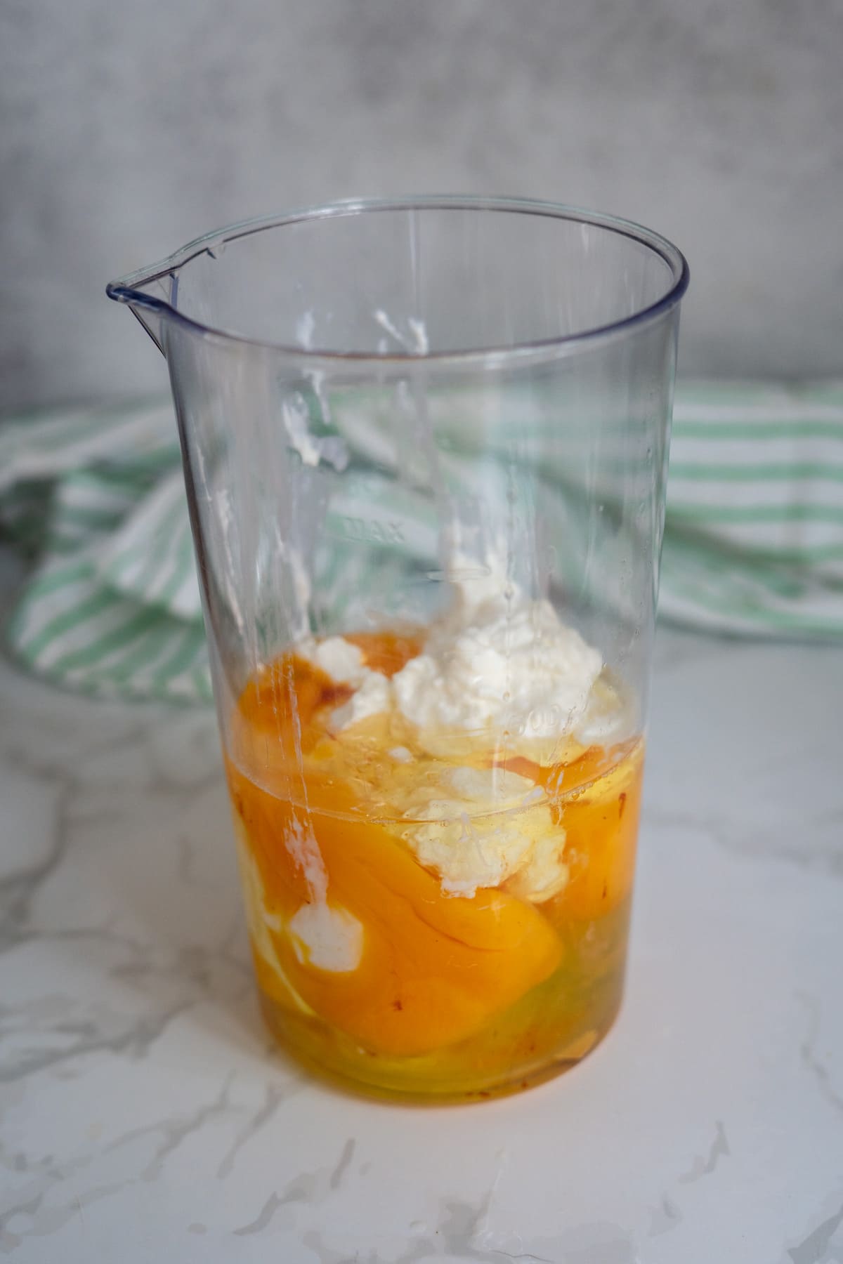 A clear plastic measuring jug containing cracked eggs and dollops of white cream or yoghurt—perfect for making egg bites for dogs—sits on a marble worktop with a striped tea towel in the background.