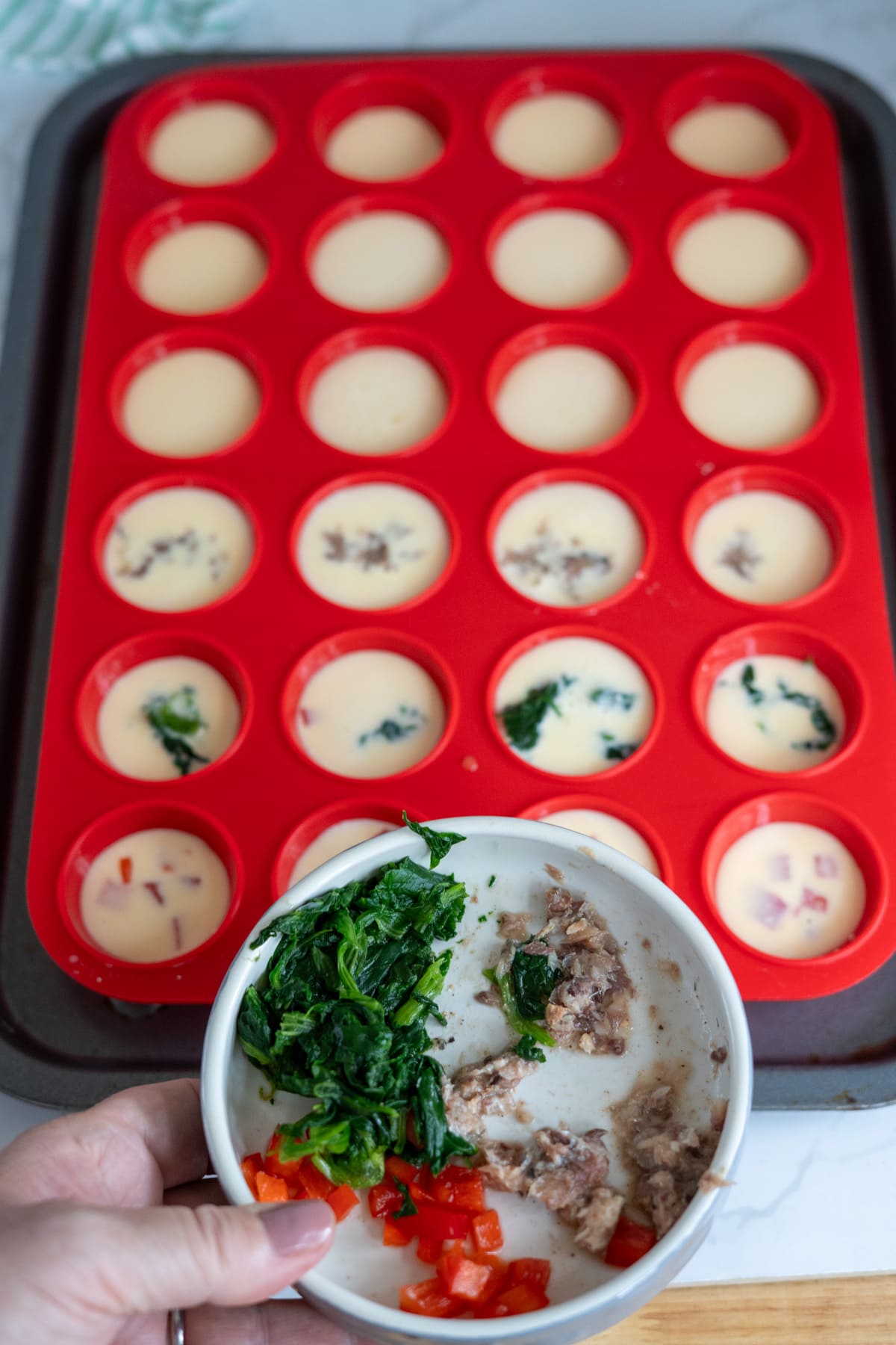 A hand holds a bowl with spinach, minced meat, and red pepper above a tray of silicone muffin moulds filled with egg mixture—some containing added fillings—preparing delicious homemade egg bites for dogs.