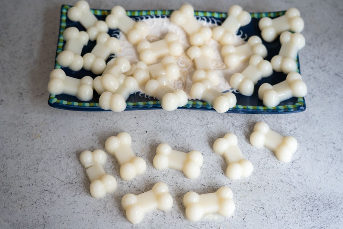 A rectangular plate holds several white, bone-shaped goat’s milk and bone broth gummies for dogs, with a few more treats scattered on a grey surface nearby.