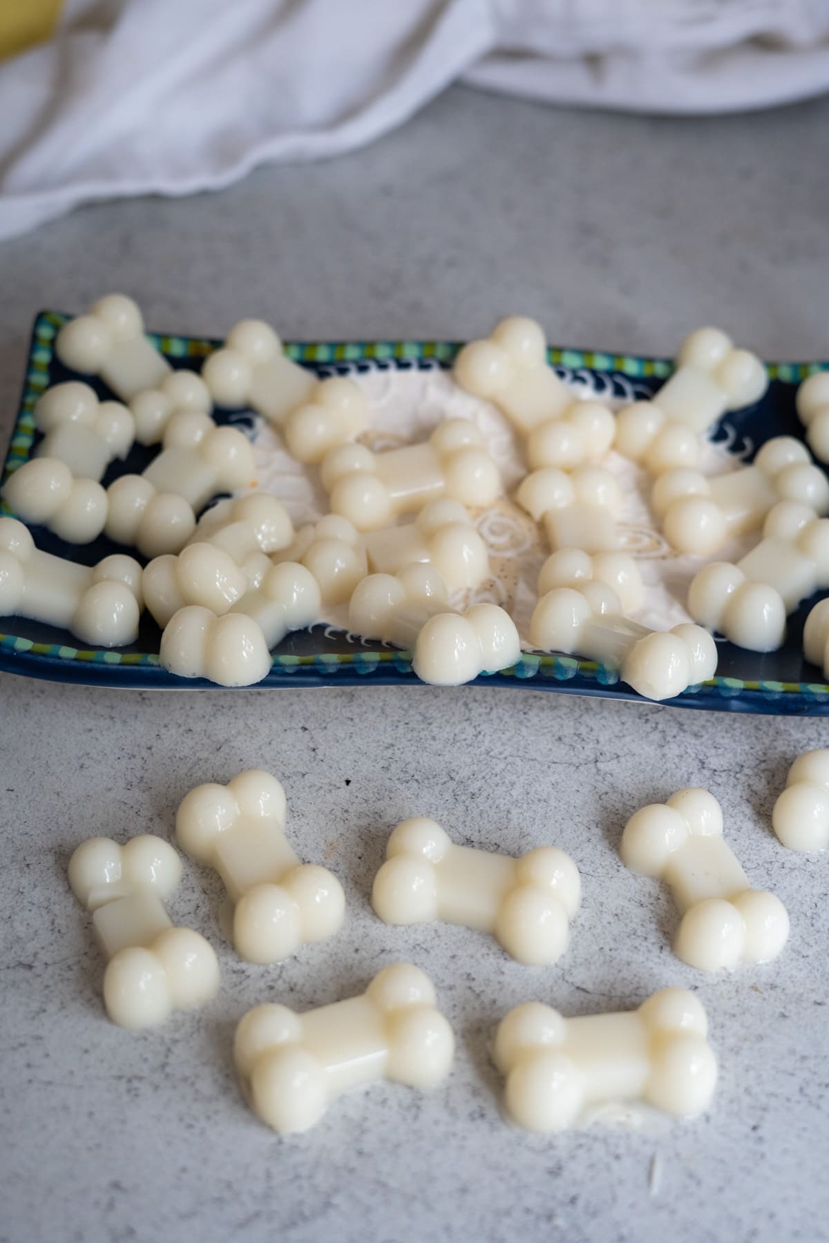 White, bone-shaped treats made from goat’s milk and bone broth gummies for dogs are arranged on a blue plate and scattered on a grey countertop, with a white cloth in the background.