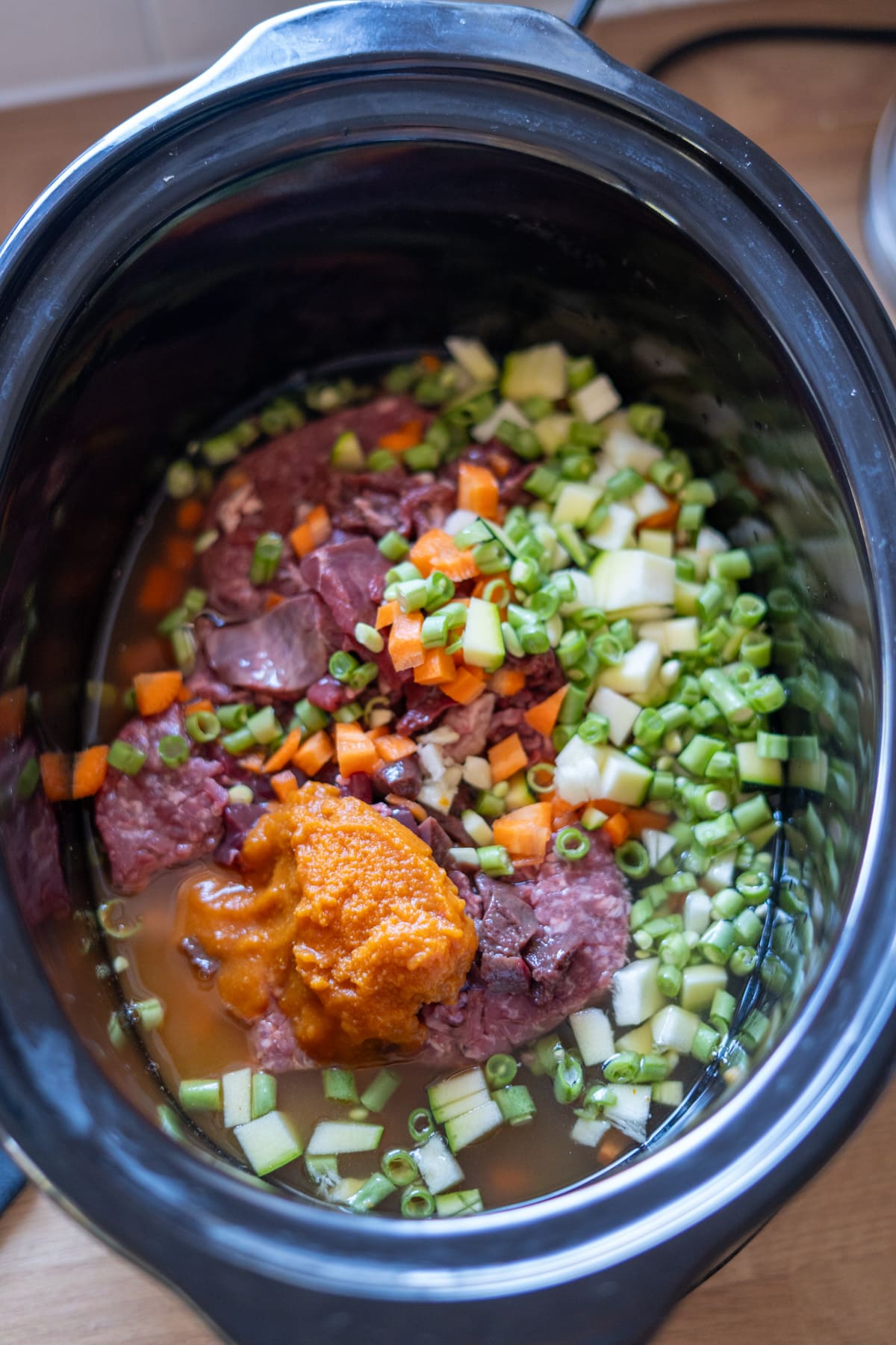 Chopped vegetables, raw meat, and orange purée are in a slow cooker with liquid, ready to be cooked into a nutritious Slow Cooker Beef Dog Food.