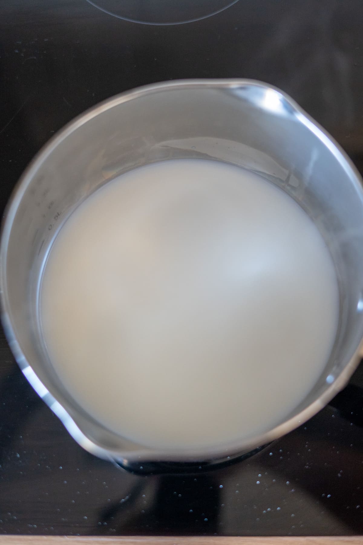 A stainless steel saucepan containing a cloudy white liquid, likely rice being cooked in water with the addition of goat’s milk, sits on a black hob.