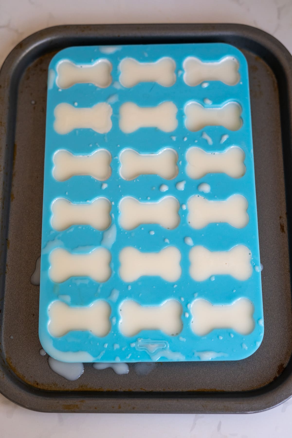 A blue silicone mould shaped like dog bones is filled with a white liquid—perfect for making goat’s milk and bone broth gummies for dogs—and placed on a metal tray, with some liquid spilt around the mould.