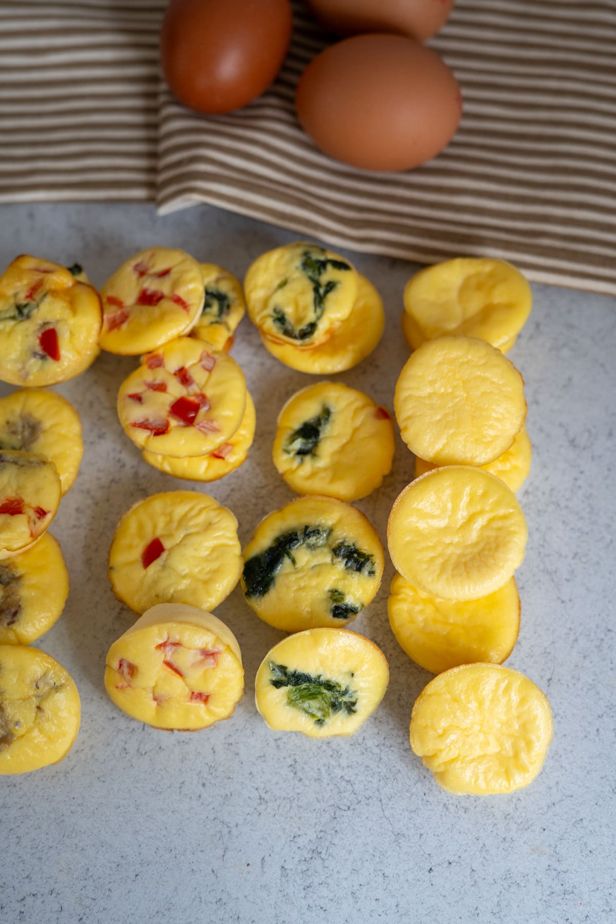 Mini egg muffins with assorted fillings are arranged on a light surface next to three whole eggs and a striped cloth in the background, creating a scene reminiscent of homemade egg bites for dogs.