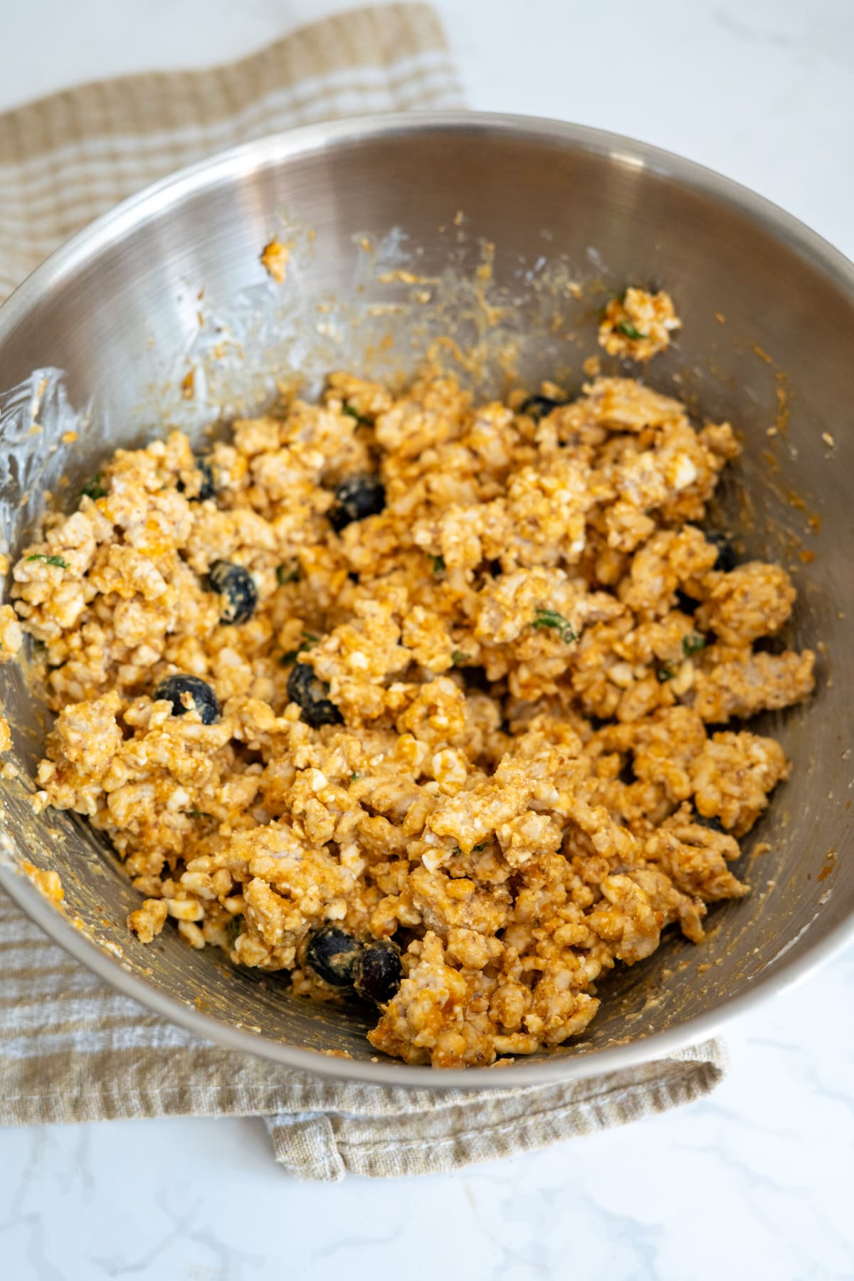 A metal mixing bowl contains a blended mixture of rice, black olives, cheese, and a reddish sauce, placed on a beige kitchen tea towel on a white marble surface.