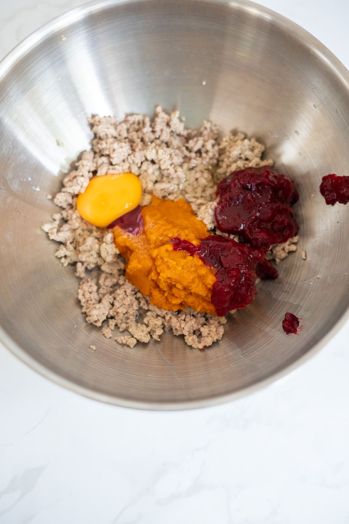 A metal bowl contains minced meat, a raw egg yolk, pumpkin purée, and cranberry sauce—perfect ingredients for homemade Turkey Pumpkin Cranberry Dog Treats.