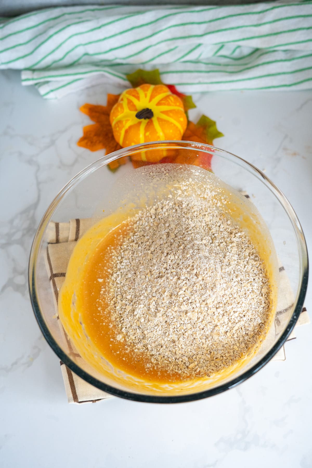 A glass bowl with a mixture of oats and pumpkin purée sits on a cloth, ready to be transformed into homemade goat milk pumpkin dog biscuits, with a small decorative pumpkin and autumn leaves in the background.