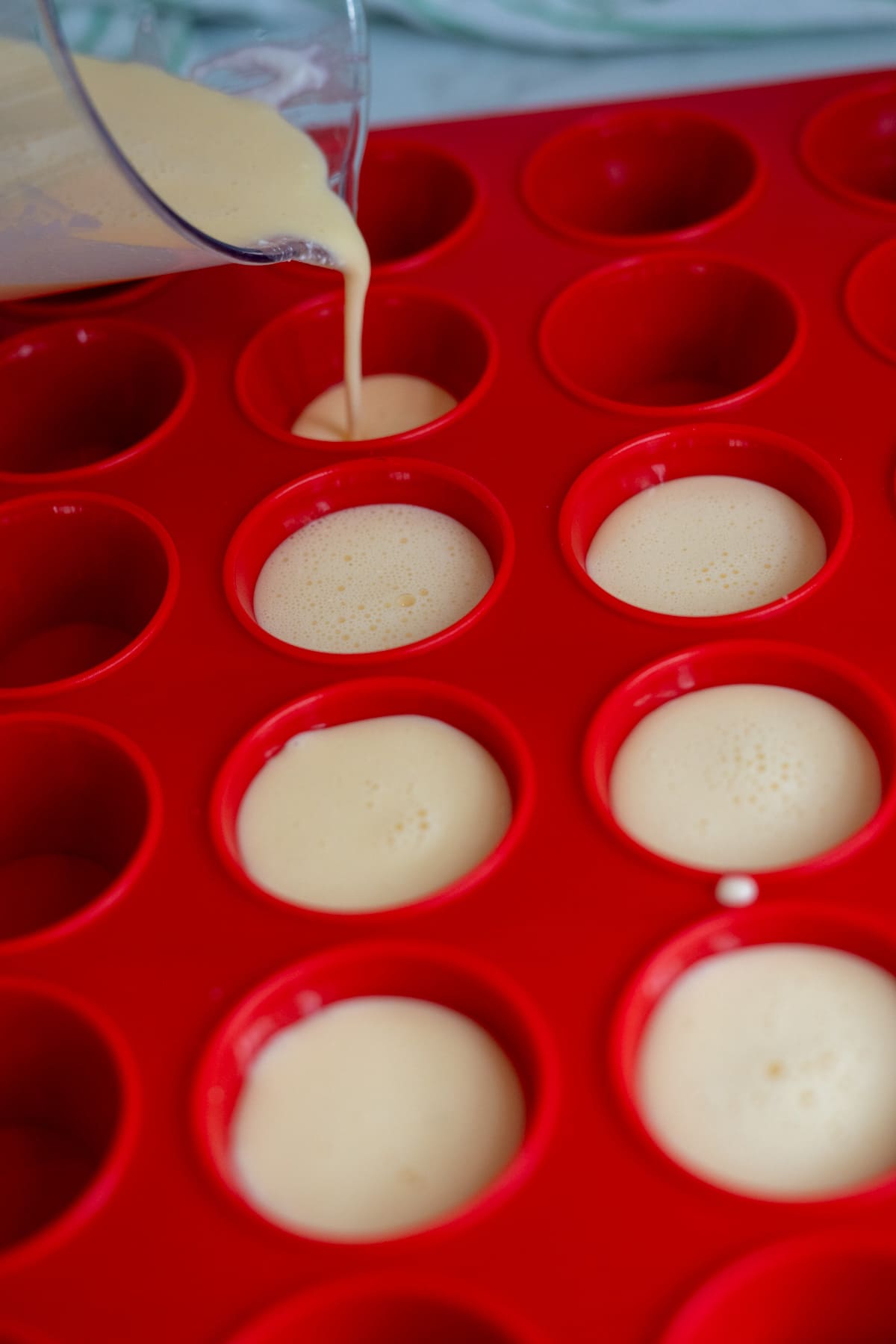 Batter is being poured from a measuring jug into a red silicone muffin tray with several wells partially filled, perfect for making homemade egg bites for dogs.